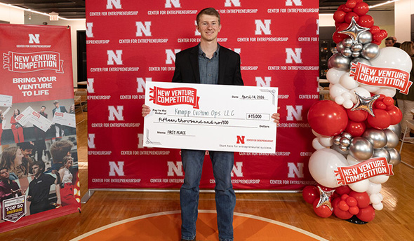 Man holding a large check at an event with red-themed banners and balloons in the background.